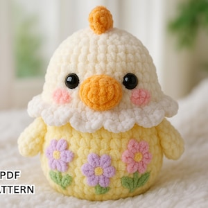 Puede incluir: Un juguete amigurumi de ganchillo en forma de pollito con un cuerpo amarillo, un cuello blanco y esponjoso y detalles de flores rosas y moradas. El pollito tiene un pequeño pico naranja y ojos negros.