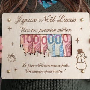 Peut inclure: Une plaque en bois avec l'inscription "Joyeux Noël Lucas" et "Voici ton premier million". La plaque comporte une ouverture rectangulaire affichant des billets en euros pour un total de 100 000. Un bonhomme de neige et un coffret cadeau sont gravés.