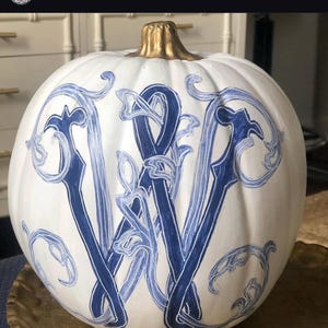 May include: A white pumpkin featuring a hand-painted dark blue letter W in a decorative, swirling script style. The pumpkin has a metallic gold-painted stem and is adorned with intricate blue filigree patterns surrounding the central letter.