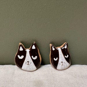 Könnte beinhalten: Zwei Keramik-Katzenkopf-Ornamente in Schwarz und Weiß bemalt. Die Katzen haben geschlossene Augen und eine weiße Nase.