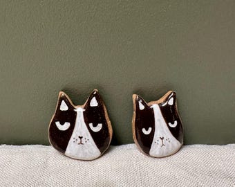 May include: Two ceramic cat head ornaments with black and white paint. The cats have closed eyes and a white nose.