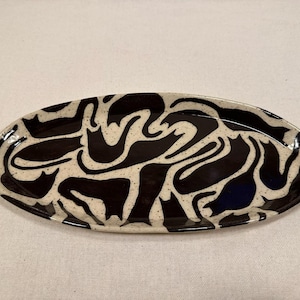 Könnte beinhalten: Ovale Keramik-Servierplatte mit einem abstrakten Design in Schwarz und Creme. Die Platte hat eine glänzende Oberfläche und einen gesprenkelten cremefarbenen Hintergrund. Das schwarze Design ist ein sich wiederholendes Muster aus geschwungenen Formen.