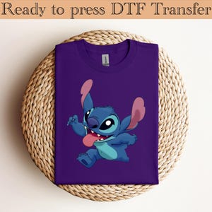 May include: A purple t-shirt featuring a cartoon character, Stitch, with blue and pink accents. The character is smiling with its tongue out. The text "Ready to press DTF Transfer" is at the top.