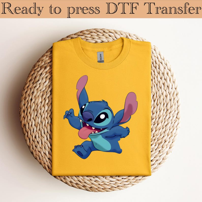 May include: A yellow t-shirt featuring a cartoon character, Stitch, with blue fur, pink ears, and a red tongue. The text "Ready to press DTF Transfer" is at the top. The shirt is on a woven, circular surface.