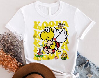 May include: White t-shirt featuring a yellow Koopa Troopa character from the Super Mario Bros. video game series. The character is flying with wings and surrounded by coins, stars, and mushrooms.