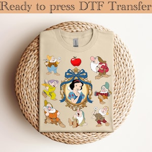 以下が含まれることがあります： 白雪姫と七人の小人のデザインが施されたベージュ色のTシャツ。デザインには白雪姫の肖像画、赤いリンゴ、様々なポーズの七人の小人が含まれています。「Ready to press DTF Transfer」の文字が上部にあります。