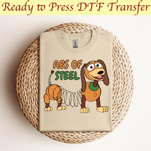 Può includere: Una maglietta beige con un'immagine di un cartone animato di un cane marrone con una molla per la coda. Il testo "ABS OF STEEL" è stampato sulla maglietta.