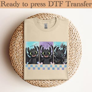 May include: Beige t-shirt featuring a colorful graphic of three cartoon dragons. The dragons are black with green eyes and are set against a backdrop of blue and purple. The shirt has the text "Ready to press DTF Transfer" at the top.