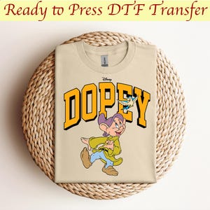 May include: A beige t-shirt with a yellow graphic of Dopey from Disney's Snow White and the Seven Dwarfs. The graphic includes the word "Dopey" in yellow letters and a small blue bird.