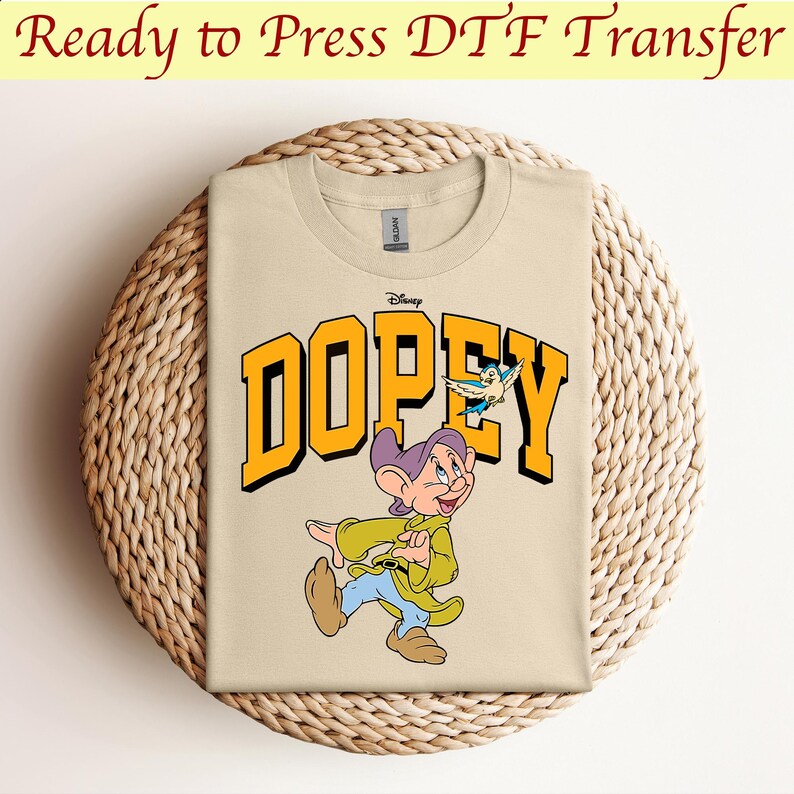 May include: A beige t-shirt with a yellow graphic of Dopey from Disney's Snow White and the Seven Dwarfs. The graphic includes the word "Dopey" in yellow letters and a small blue bird.