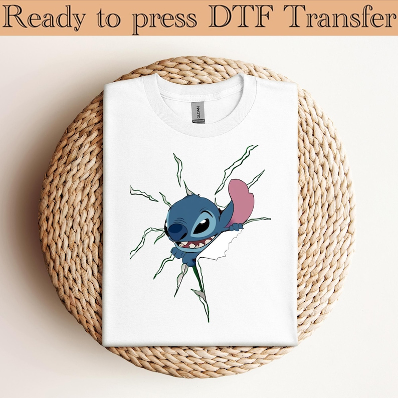 K&ouml;nnte beinhalten: Wei&szlig;es T-Shirt mit einem blauen Cartoon-Charakter-Design, mit einem rosa Ohr, das durch einen zerrissenen Bereich bricht. Der Text "Ready to press DTF Transfer" steht oben. Das T-Shirt wird auf einer gewebten, kreisf&ouml;rmigen Oberfl&auml;che pr&auml;sentiert.