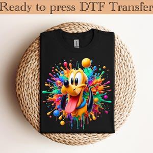 Könnte beinhalten: Schwarzes T-Shirt mit einem farbenfrohen Cartoon-Bild von Pluto und einem Farbspritzer-Design. Der Text "Ready to press DTF Transfer" steht oben. Das Shirt liegt gefaltet auf einer gewebten, kreisförmigen Oberfläche.