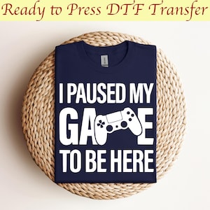 May include: A navy blue t-shirt with white text that reads "I paused my game to be here". A white video game controller graphic is centered between the words "game" and "to".