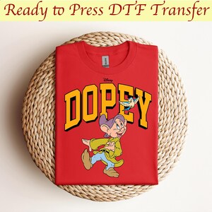May include: Red t-shirt with a yellow "DOPEY" graphic featuring a cartoon image of Dopey from Disney's Snow White and the Seven Dwarfs. The graphic also includes a small blue bird.
