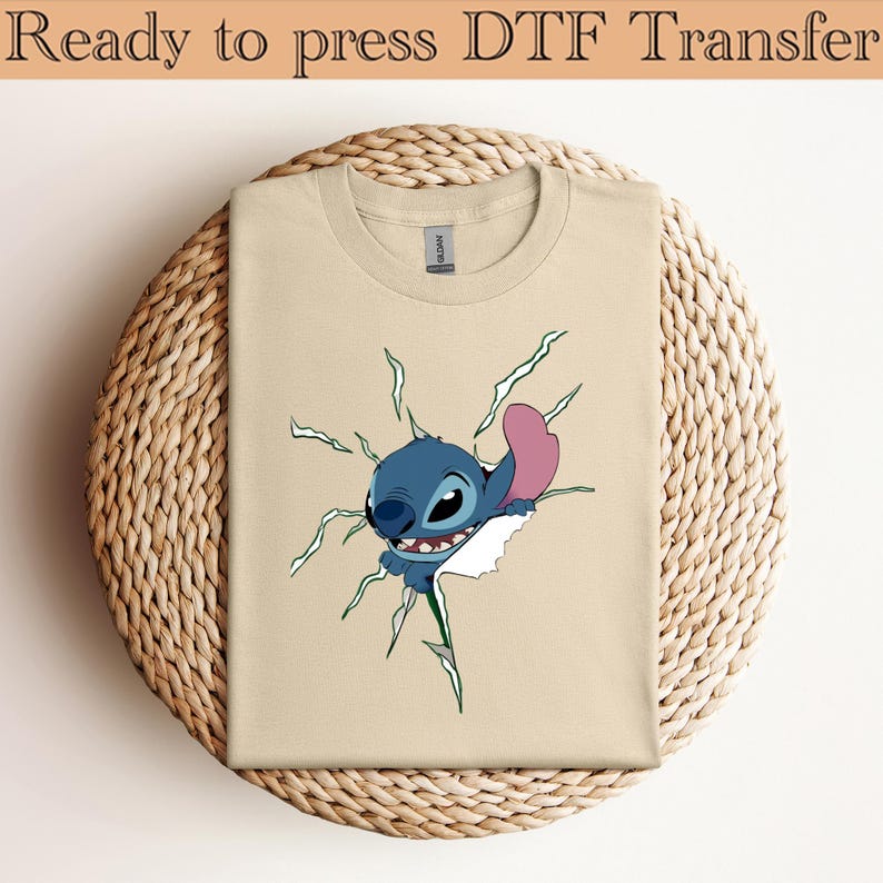 K&ouml;nnte beinhalten: Beiges T-Shirt mit einem Cartoon-Charakter, Stitch, der durch ein Loch bricht. Stitch ist blau mit einem rosa Ohr und einem breiten Grinsen. Auf dem T-Shirt steht oben der Text "Ready to press DTF Transfer".