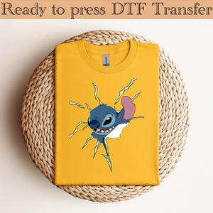 K&ouml;nnte beinhalten: Ein gelbes T-Shirt mit einem Cartoon-Charakter, der aus dem Stoff bricht. Der Charakter ist blau mit einem rosa Ohr. Der Text "Ready to press DTF Transfer" befindet sich oben.