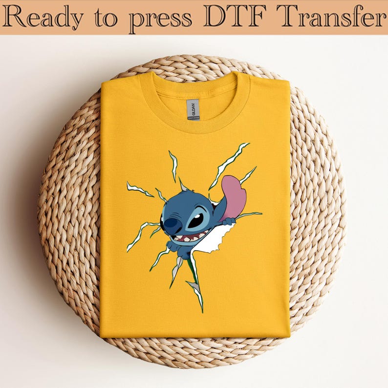 K&ouml;nnte beinhalten: Ein gelbes T-Shirt mit einem Cartoon-Charakter, der aus dem Stoff bricht. Der Charakter ist blau mit einem rosa Ohr. Der Text "Ready to press DTF Transfer" befindet sich oben.