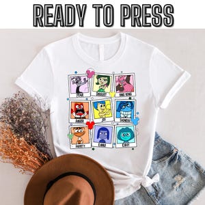 以下が含まれることがあります： 映画「インサイド・ヘッド」のキャラクターが描かれた白いTシャツ。各キャラクターは、カラフルなポラロイドフレームの中にあり、その下に名前が書かれています。キャラクターは、喜び、悲しみ、怒り、恐怖、嫌悪、ビング・ボン、不安、エミュ、そして羨望です。