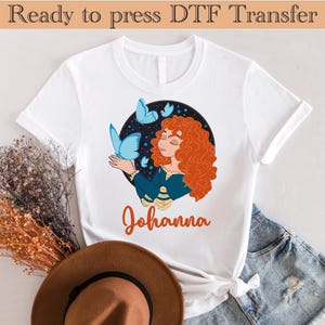 以下が含まれることがあります： 赤い髪と青い蝶を持つ女性のグラフィックが描かれた白いTシャツ。「Johanna」という名前がオレンジ色の筆記体で画像の下に書かれています。「Ready to press DTF Transfer」というテキストが上部にあります。