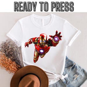 May include: White t-shirt with a red and gold Iron Man graphic. The graphic shows Iron Man flying with his hand outstretched, firing a repulsor beam. The text "READY TO PRESS" is at the top of the image.
