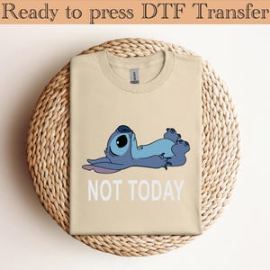 K&ouml;nnte beinhalten: Ein beiges T-Shirt mit einer Grafik eines blauen Alien-Charakters, der auf dem R&uuml;cken liegt, mit dem darunter in Wei&szlig; gedruckten Text "NOT TODAY". Der obere Teil des Bildes enth&auml;lt den Text "Ready to press DTF Transfer" in Dunkelbraun.