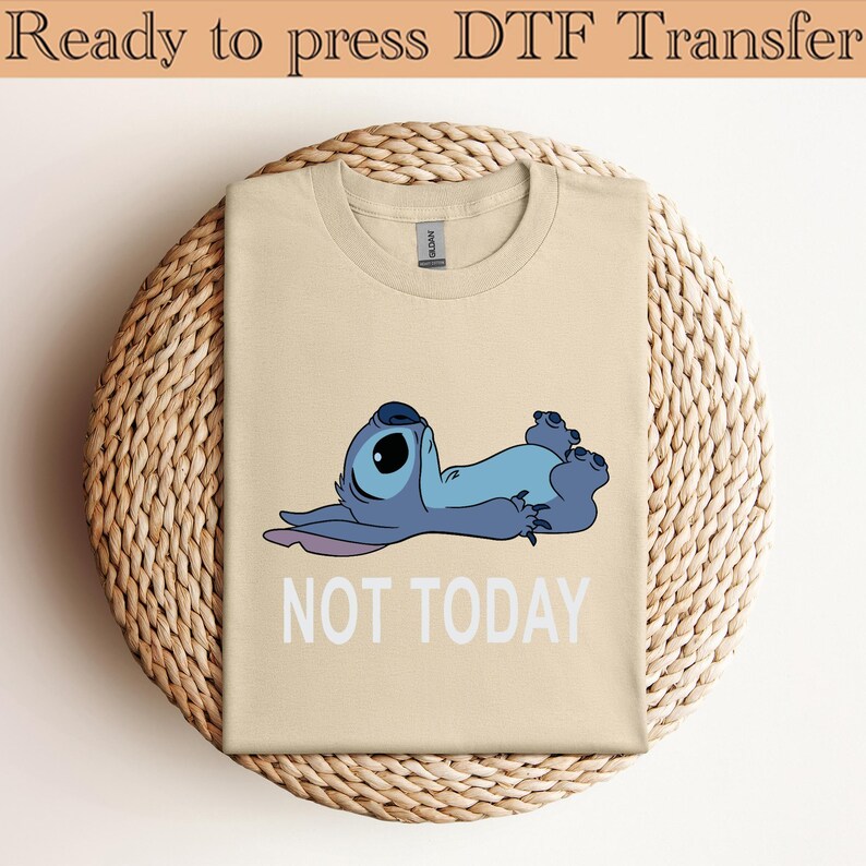 K&ouml;nnte beinhalten: Ein beiges T-Shirt mit einer Grafik eines blauen Alien-Charakters, der auf dem R&uuml;cken liegt, mit dem darunter in Wei&szlig; gedruckten Text "NOT TODAY". Der obere Teil des Bildes enth&auml;lt den Text "Ready to press DTF Transfer" in Dunkelbraun.