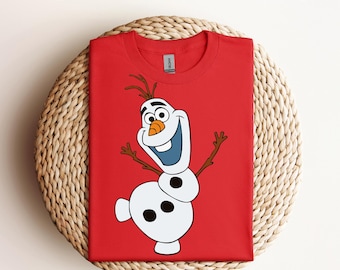 Puede incluir: Camiseta roja con un gráfico de Olaf de Frozen, un personaje de dibujos animados blanco. La camiseta está doblada y colocada sobre una superficie de mimbre tejido.