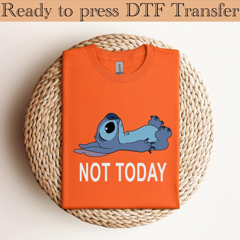 K&ouml;nnte beinhalten: Ein leuchtend orangefarbenes T-Shirt mit Rundhalsausschnitt und einer Grafik eines blauen Alien-Charakters, der auf dem R&uuml;cken liegt, mit dem darunter gedruckten wei&szlig;en Text NOT TODAY. Das Shirt hat ein klassisches Kurzarmdesign und ein Etikett.