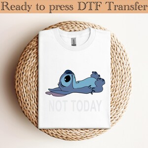 K&ouml;nnte beinhalten: Ein wei&szlig;es T-Shirt mit einer Grafik eines blauen Alien-Charakters, der auf dem R&uuml;cken liegt und die Augen geschlossen hat. Unter der Figur steht der Text NOT TODAY in hellgrauen Gro&szlig;buchstaben. Das T-Shirt hat einen Rundhalsausschnitt.