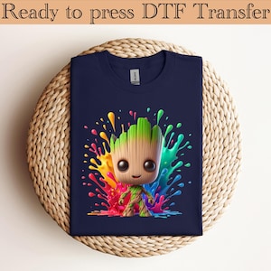 May include: A navy blue t-shirt featuring a graphic of a small, tree-like character with large black eyes and a wood-textured body. The character is surrounded by a vibrant, multi-colored paint splatter design in shades of red, yellow, green, and blue.