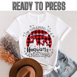 Puede incluir: Camiseta blanca con un diseño navideño. El diseño presenta un círculo a cuadros rojos y negros con una silueta de castillo, renos y el texto "I'm Dreaming of a Hogwarts Christmas". Las palabras "READY TO PRESS" están en la parte superior.