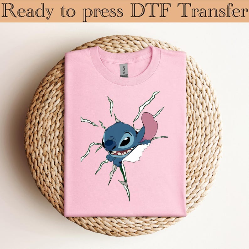 K&ouml;nnte beinhalten: Ein rosa T-Shirt mit einem Cartoon-Stitch-Design, das durch ein zerrissenes Design bricht. Die blaue Figur hat einen schelmischen Blick. Der Text "Ready to press DTF Transfer" befindet sich oben.