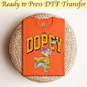May include: Orange t-shirt with a graphic of Dopey from Disney's Snow White and the Seven Dwarfs. The graphic features Dopey walking with a bird flying above him. The text "Disney" and "DOPEY" are in yellow letters.