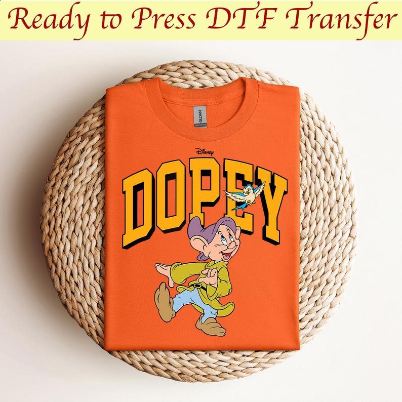 May include: Orange t-shirt with a graphic of Dopey from Disney's Snow White and the Seven Dwarfs. The graphic features Dopey walking with a bird flying above him. The text "Disney" and "DOPEY" are in yellow letters.