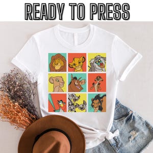Puede incluir: Camiseta blanca con un gráfico colorido de personajes de la película El Rey León. El gráfico presenta a Simba, Nala, Pumbaa, Timon, Scar y Mufasa en un patrón de cuadrícula. El texto "READY TO PRESS" está en la parte superior de la imagen.