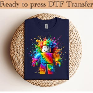 Puede incluir: Camiseta azul marino con un gráfico colorido de una figura de bloques con una cara sonriente y salpicaduras de pintura arcoíris. El texto "Ready to press DTF Transfer" está en la parte superior.