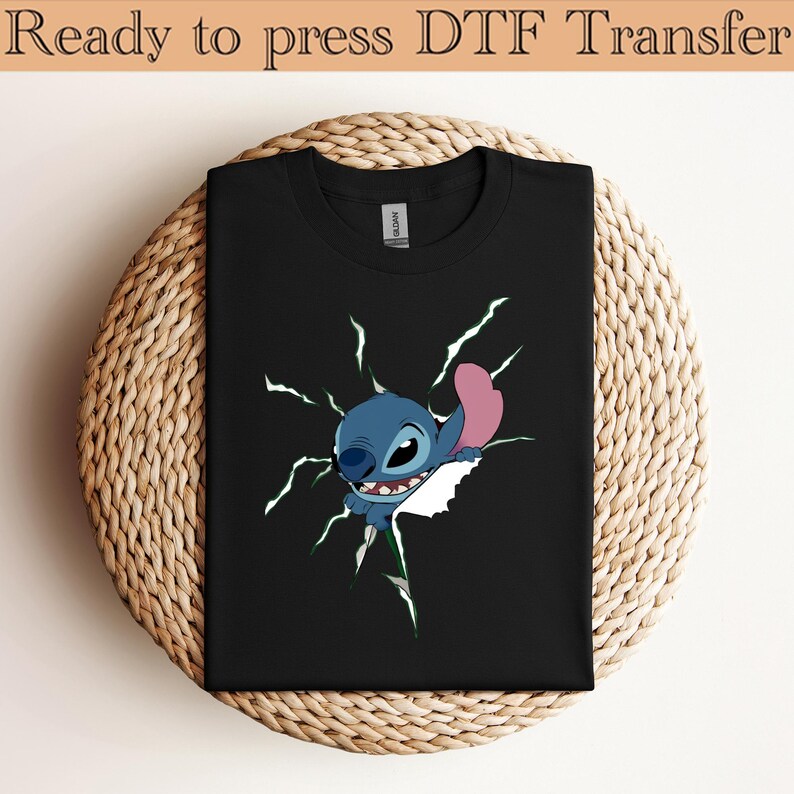 K&ouml;nnte beinhalten: Schwarzes T-Shirt mit einem Cartoon-Charakter, der durch eine zerrissene wei&szlig;e Oberfl&auml;che bricht. Der Charakter ist blau mit einem rosa Ohr und einem breiten Grinsen. Der Text "Ready to press DTF Transfer" steht oben.