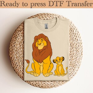 May include: A beige t-shirt with a DTF transfer of Simba and Mufasa from The Lion King. The transfer is a cartoon image of the two lions sitting side by side.