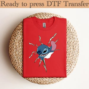 K&ouml;nnte beinhalten: Ein rotes T-Shirt mit einem Cartoon-Stitch-Design, das durch einen zerrissenen Effekt bricht. Der Text "Ready to press DTF Transfer" befindet sich oben. Das T-Shirt wird auf einer kreisf&ouml;rmigen Webmatte pr&auml;sentiert.