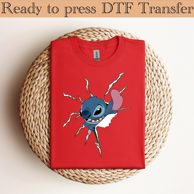 K&ouml;nnte beinhalten: Ein rotes T-Shirt mit einem Cartoon-Stitch-Design, das durch einen zerrissenen Effekt bricht. Der Text "Ready to press DTF Transfer" befindet sich oben. Das T-Shirt wird auf einer kreisf&ouml;rmigen Webmatte pr&auml;sentiert.
