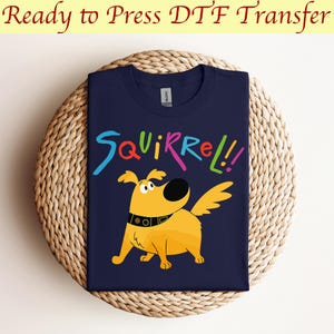 May include: A navy blue t-shirt featuring a yellow cartoon dog and the text "Squirrel!" in colourful letters. The shirt is ready to be pressed with a DTF transfer.