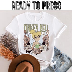 Könnte beinhalten: Weißes T-Shirt mit einem Grafikdruck von Tinkerbell mit Blumen und dem Text "Tinker Bell".