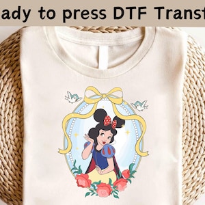 以下が含まれることがあります： クリーム色のTシャツ。DTF転写で、バラと鳥の装飾フレームに入った白雪姫が描かれています。「Ready to press DTF Transfer」の文字が画像上部にあります。