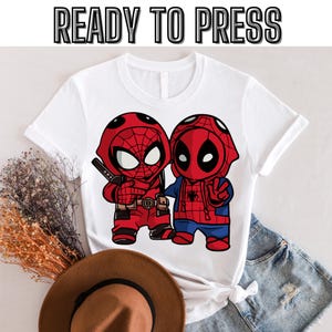 May include: White t-shirt with a graphic print of two cartoon superheroes, Spiderman and Deadpool, in red and blue colors. The text "READY TO PRESS" is printed at the top of the image.