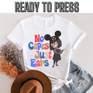 May include: White t-shirt with a cartoon image of a woman wearing a black dress and Mickey Mouse ears. The text "No capes just ears" is printed in colorful letters on the shirt.