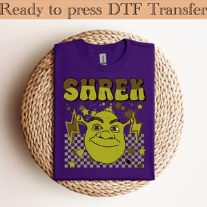 May include: A purple t-shirt featuring the word "SHREK" in yellow and brown lettering, with a cartoon image of Shrek below. The design includes lightning bolts, stars, and a checkered pattern. The text "Ready to press DTF Transfer" is at the top.