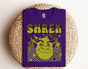 May include: A purple t-shirt featuring the word "SHREK" in yellow and brown lettering, with a cartoon image of Shrek below. The design includes lightning bolts, stars, and a checkered pattern. The text "Ready to press DTF Transfer" is at the top.