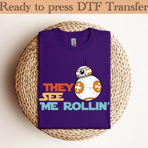 Puede incluir: Una camiseta morada con un diseño de dibujos animados de un droide BB-8 y el texto "THEY SEE ME ROLLIN'". El diseño incluye texto naranja, amarillo y azul. Las palabras "Ready to press DTF Transfer" están en la parte superior.