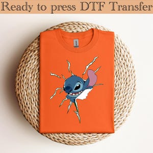 K&ouml;nnte beinhalten: Orangefarbenes T-Shirt mit einem Cartoon-Charakter, der durch eine zerrissene Oberfl&auml;che bricht. Der Charakter ist blau mit einem rosa Ohr. Der Text "Ready to press DTF Transfer" steht oben.