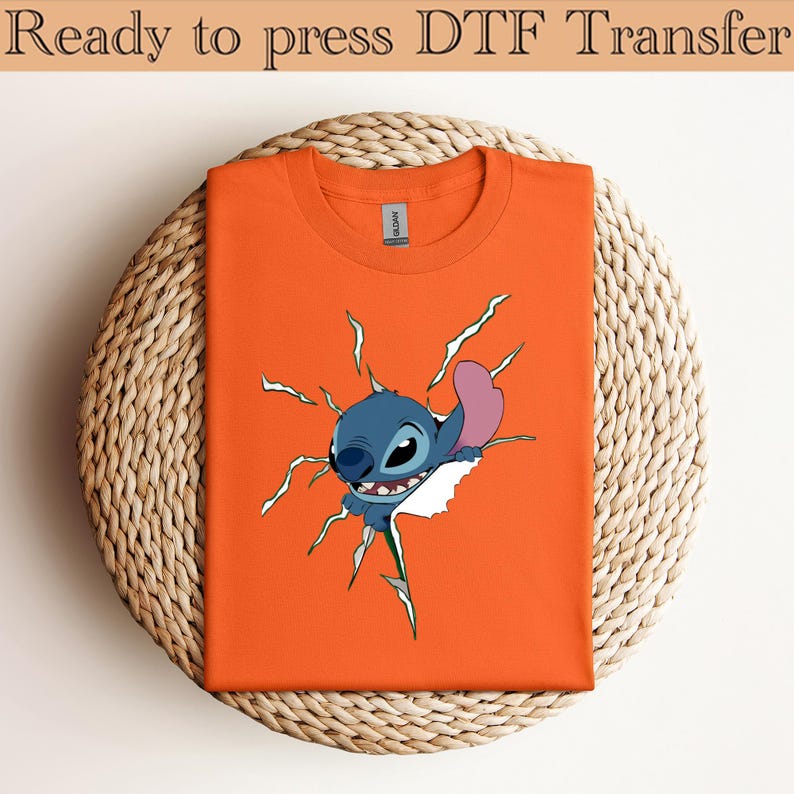 K&ouml;nnte beinhalten: Orangefarbenes T-Shirt mit einem Cartoon-Charakter, der durch eine zerrissene Oberfl&auml;che bricht. Der Charakter ist blau mit einem rosa Ohr. Der Text "Ready to press DTF Transfer" steht oben.